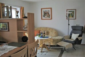 A living room with a sofa, armchair, and a small table with a lamp. There are framed pictures on the wall and a window with curtains. - House by the spa park (Bad Kohlgrub)
