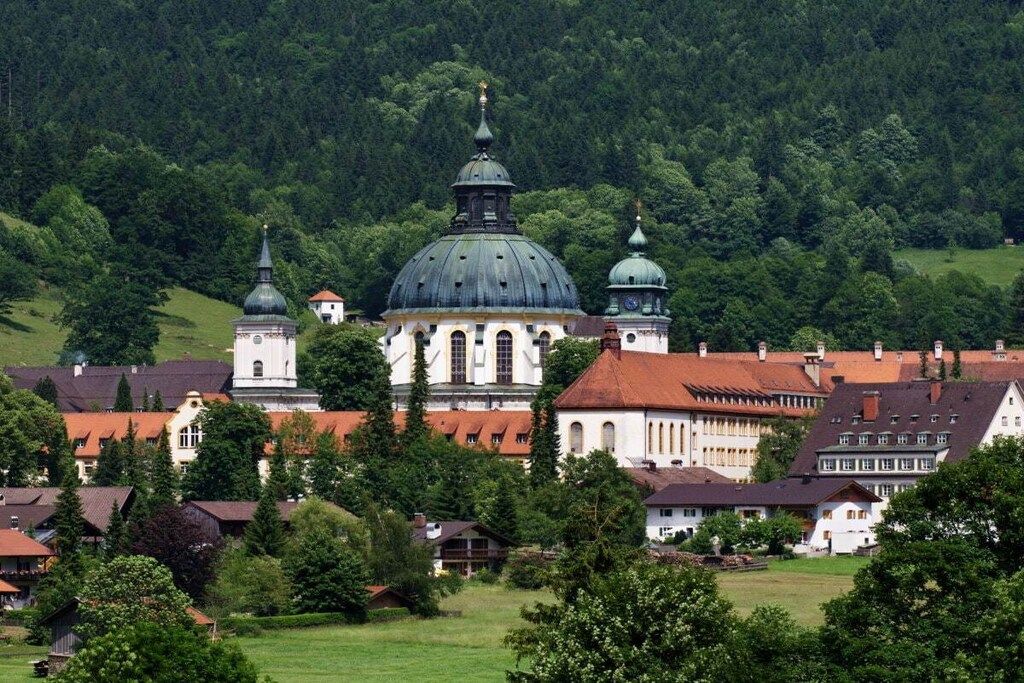 Ein historisches Gebäude mit einer großen, zentralen Kuppel und kleineren Kuppeln, umgeben von traditionellen Häusern und üppiger Vegetation.
