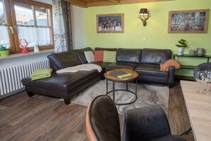 Ein Wohnzimmer mit einer Eckcouch, einem runden Couchtisch und einem Esstisch mit Stühlen. Zwei Wandlampen und ein Fenster mit Vorhängen sind vorhanden.