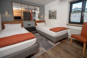 Ein Hotelzimmer mit zwei Betten, einem großen Spiegel, einem Gemälde, einem Fenster mit Blick und einem Sessel.