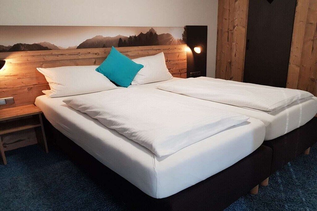 A double bed with white bedding and a blue pillow, a wooden headboard, and a nightstand with a lamp.