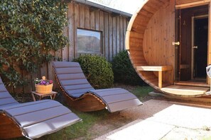 A wooden cabin with a circular exterior wall, a small window, and a door. There are two lounge chairs with cushions and a small table with a basket of flowers. - Holiday apartment with 1 bedroom (Dollerup)