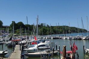 A marina with multiple boats docked, a small building, and a forested hill in the background. - Holiday apartment with 1 bedroom (Dollerup)