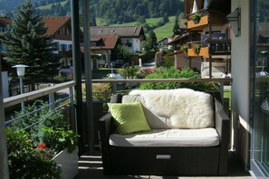 Ein Balkon mit einer Korbusitzgruppe, einem grünen Kissen und Blick auf ein Wohngebiet mit Häusern und Grünflächen.