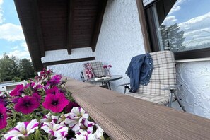 Ein überdachter Außenbereich mit einer hölzernen Bank, einem karierten Sessel und einem Tisch mit einem Buch und Blumen.