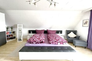 Un dormitorio con cama, mesita de noche, estantería, sofá gris y lámpara en el techo.
