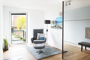 A modern living room with a grey armchair, a small ottoman, a floor lamp, and a view through a glass door to an outdoor area with plants. - Holiday apartment with 2 bedrooms (Tönning)