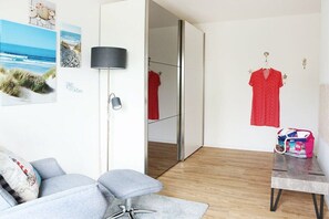 A room with a grey sofa, a small table, a lamp, a red dress on a hanger, and a bag on the table. - Holiday apartment with 2 bedrooms (Tönning)