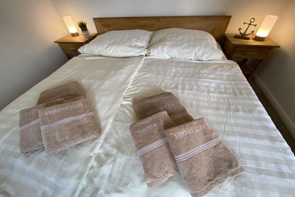 A neatly made bed with white linens and four brown towels with 'Nordstjerng' text, two bedside tables with lamps, and a wooden headboard.