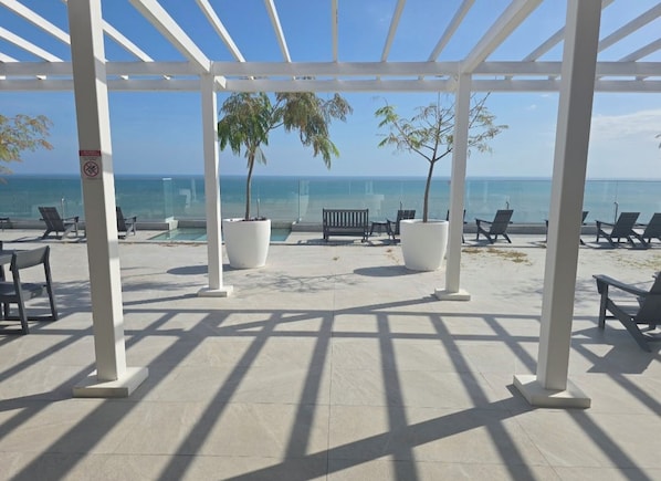 A covered outdoor area with a pool, seating, and a view of the ocean. - Luxury apartment with spectacular ocean views—right on the water, with pools, a fitness center, and a rooftop terrace (Jacó)