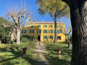 A yellow building with a symmetrical facade, a central entrance, and a gravel pathway leading to the entrance. - Charming & large 3-bedroom apartment in Provençal bastide (Grasse)