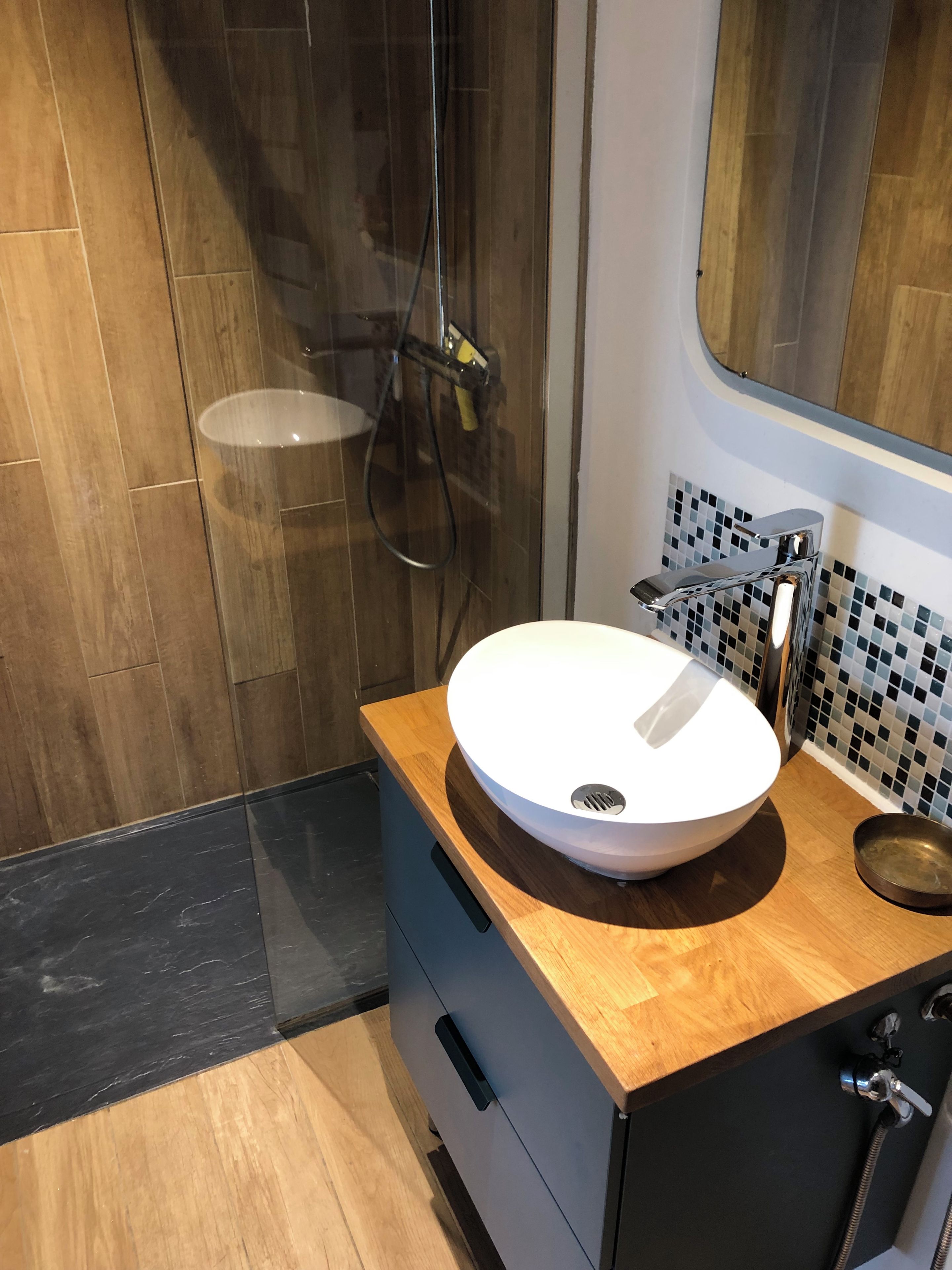 Un baño moderno con tocador de madera, un lavabo blanco, ducha con puerta de vidrio y salpicadero de azulejos.