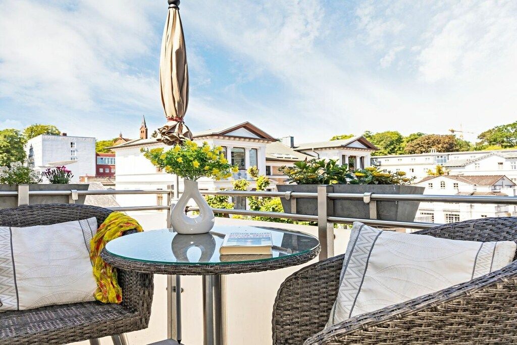 Ein Balkon mit Korbmöbeln, einem Glastisch und einer weißen Vase mit gelben Blumen.