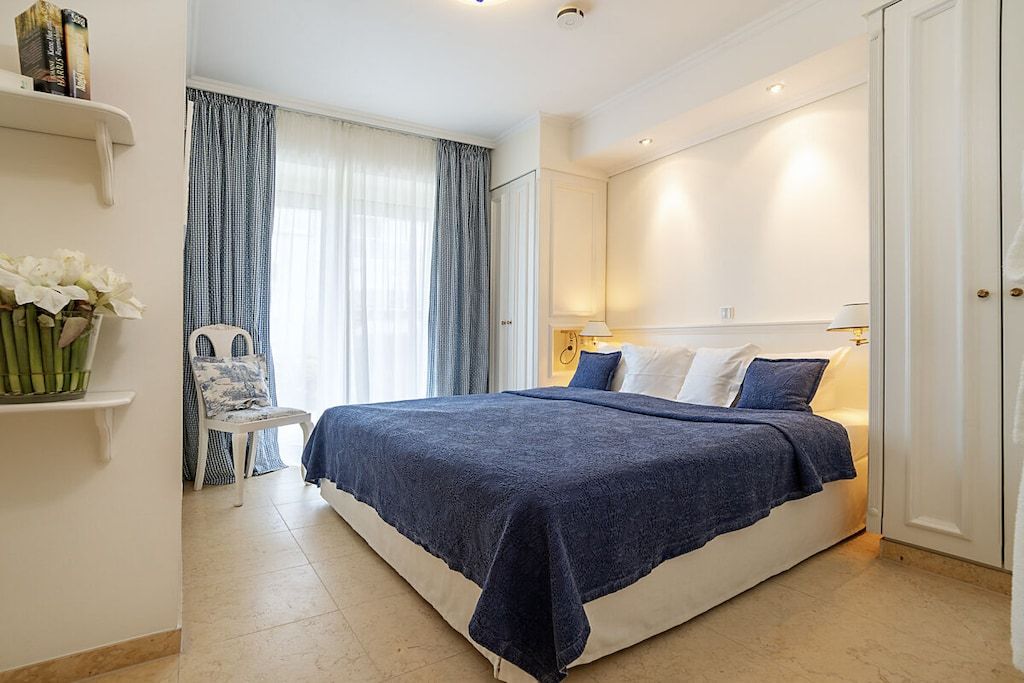 Quarto com cama grande, uma cadeira, mesa de cabeceira e janela com cortinas.