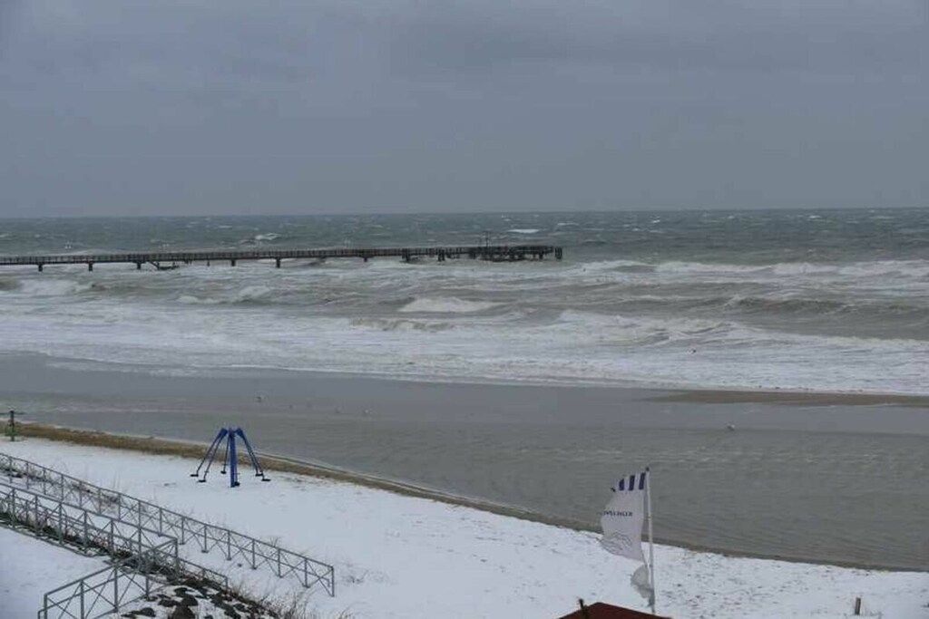 A beach with a pier extending into the water, a snow-covered area, and a flag indicating a public space.