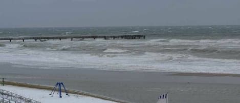 Una playa con un muelle que se adentra en el agua, una zona nevada y una bandera que indica un espacio público.