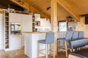 Eine kompakte Küche mit weißen Schränken, einer Mikrowelle und einem eingebauten Ofen. Zwei Hocker mit grauen Kissen sowie eine Bank mit dunklem Kissen sind vorhanden.