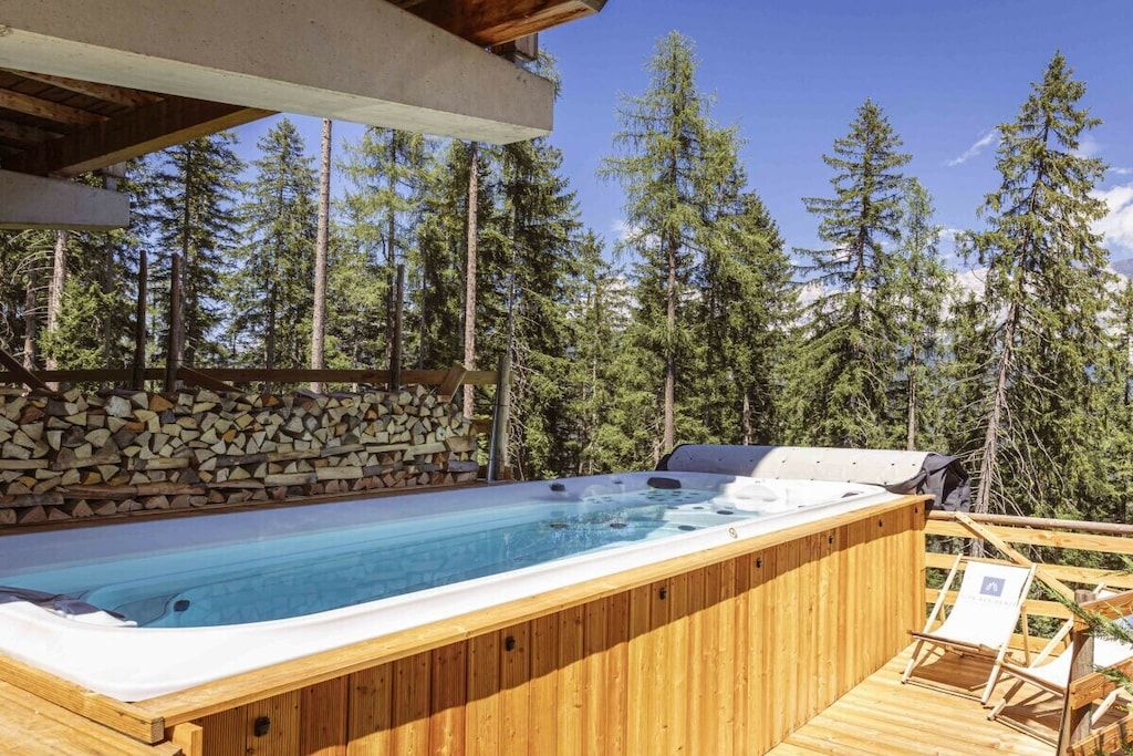 Una vasca idromassaggio su un pontile di legno, circondata da un muro in pietra e alberi alti.