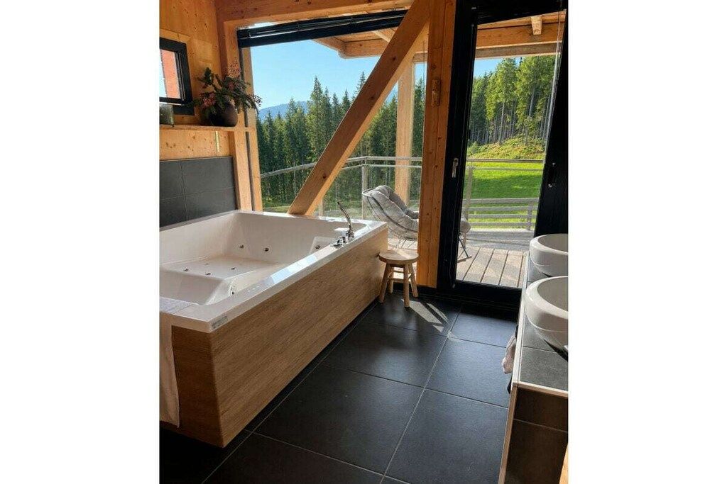 A modern bathroom with a large bathtub, a wooden bench, and a view of a forest through a glass door.