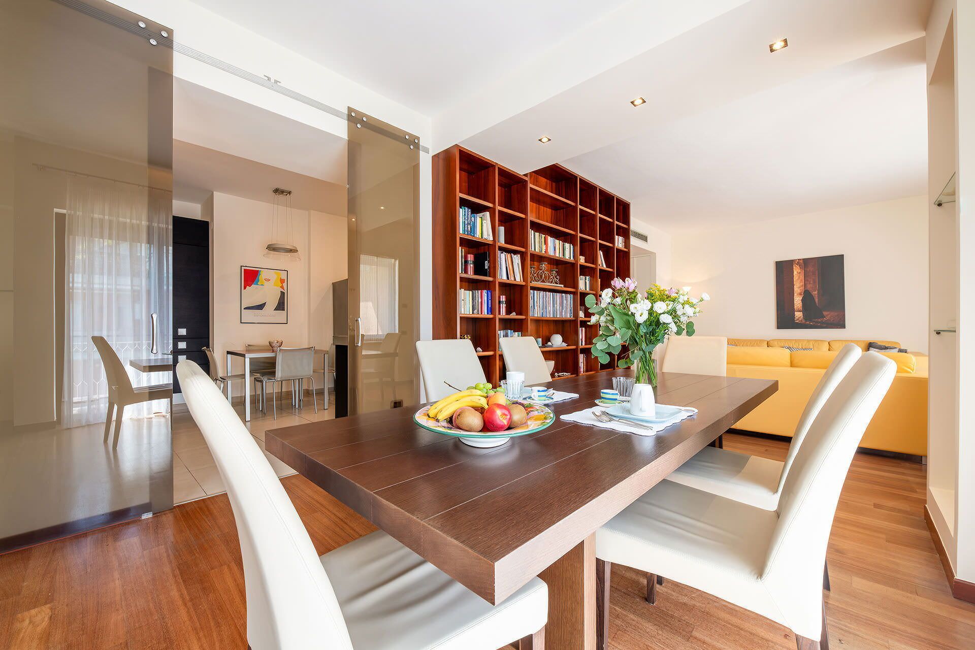 A dining area with a wooden table, white chairs, and a bookshelf filled with books.