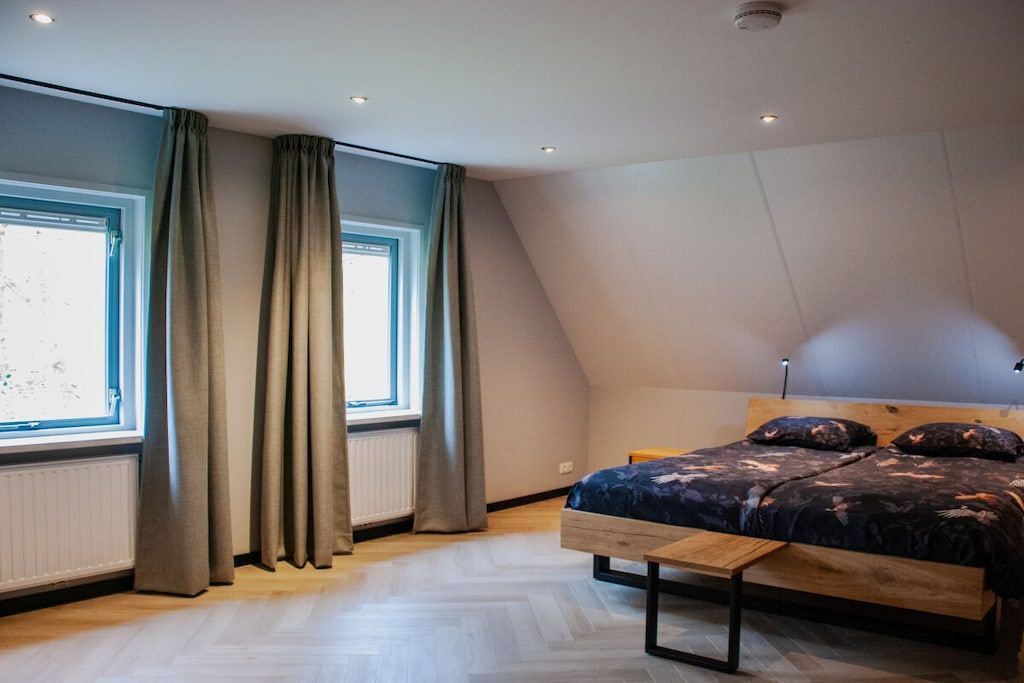 Een slaapkamer met een schuine kap, een bed met een houten hoofdbord, een bankje en een raam met gordijnen.