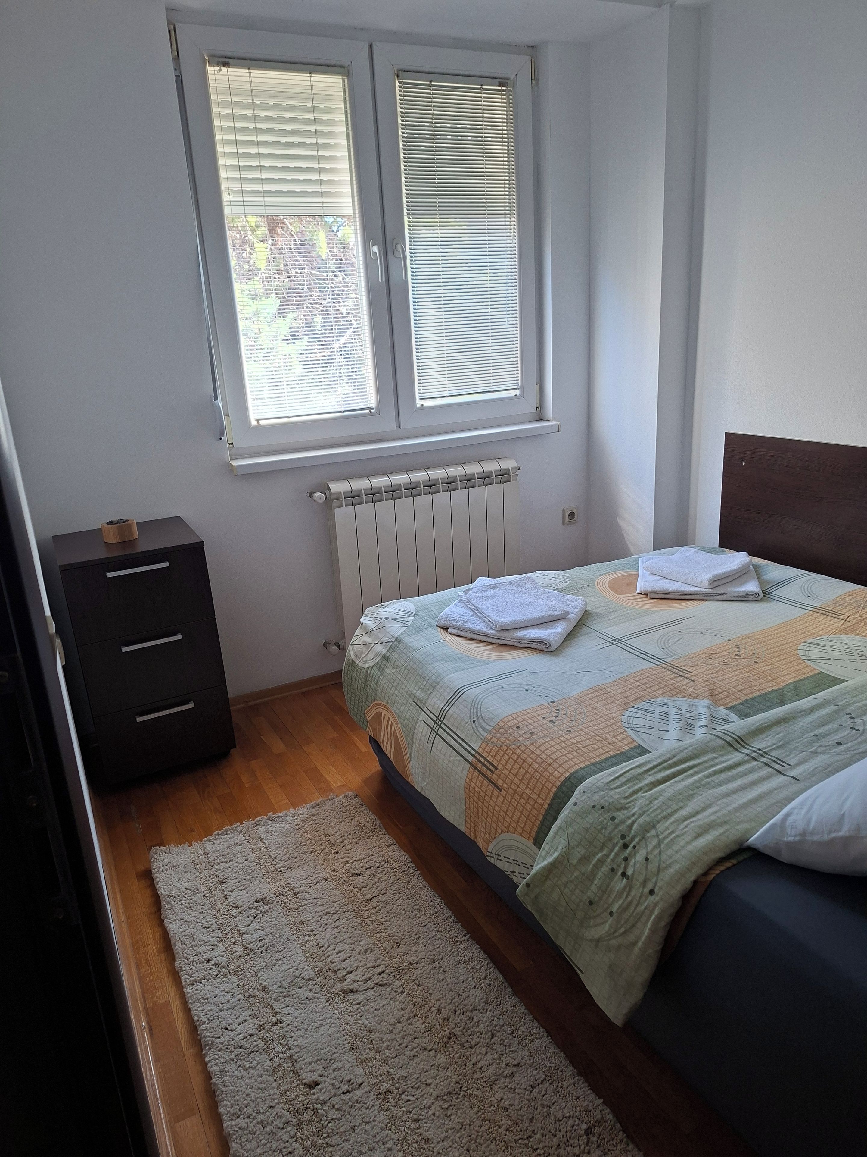 A bedroom with a bed, a radiator, a window with blinds, a dark dresser, and a carpet.