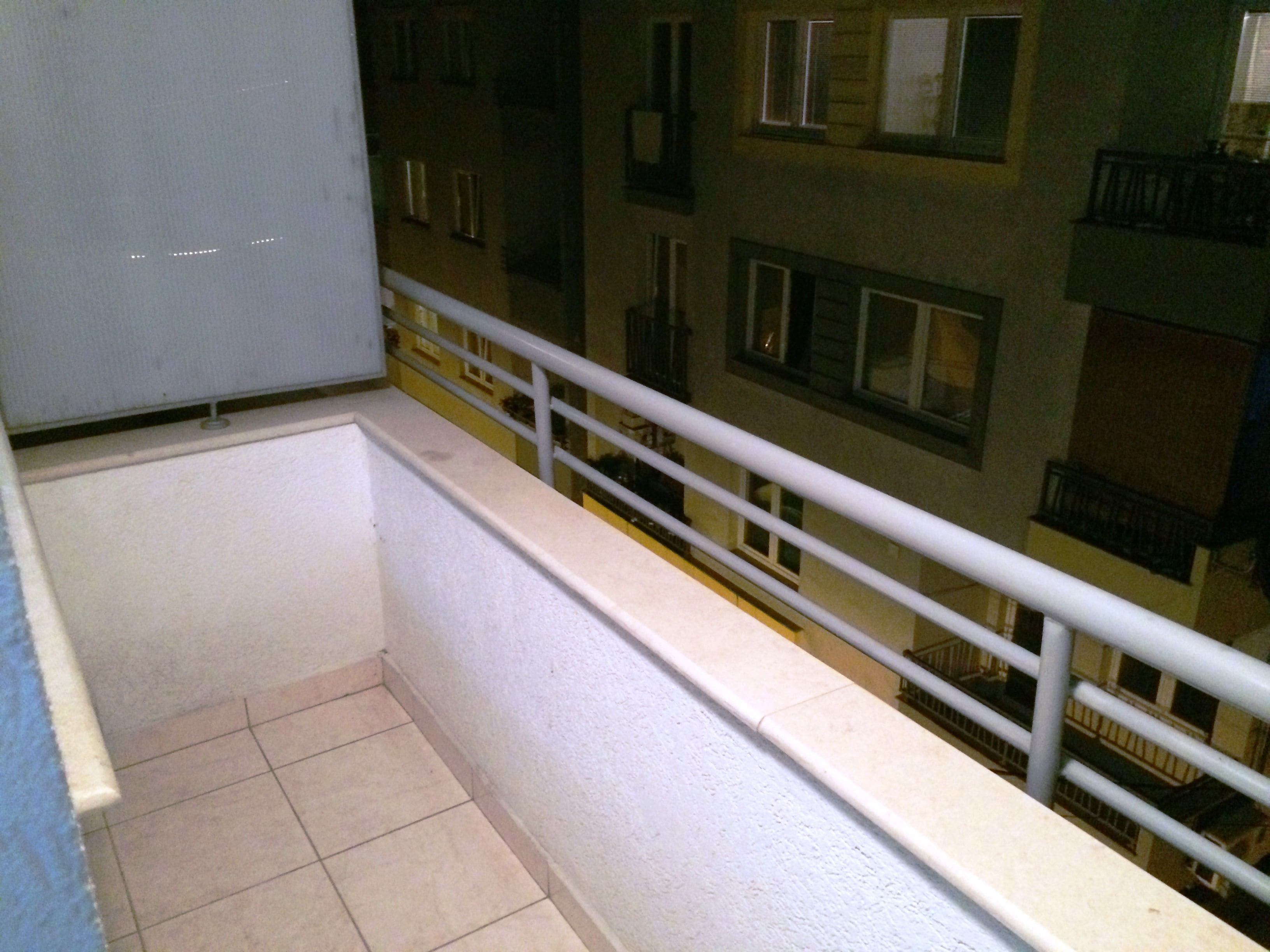 Un balcón con barandilla blanca, suelo de baldosas y vistas a un edificio con varias ventanas y balcones.