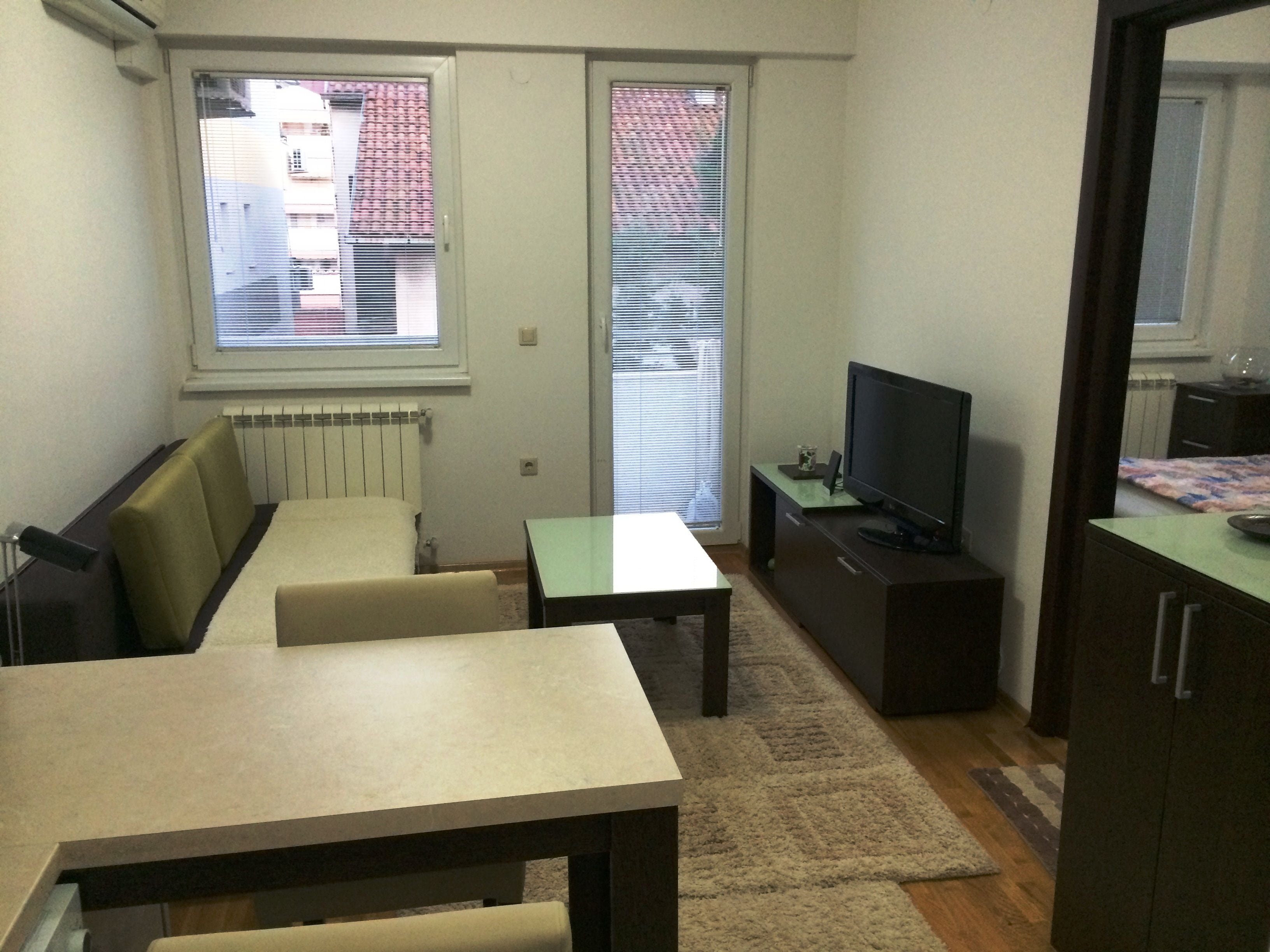 A small, furnished room with a sofa, a table, a television, and a radiator.