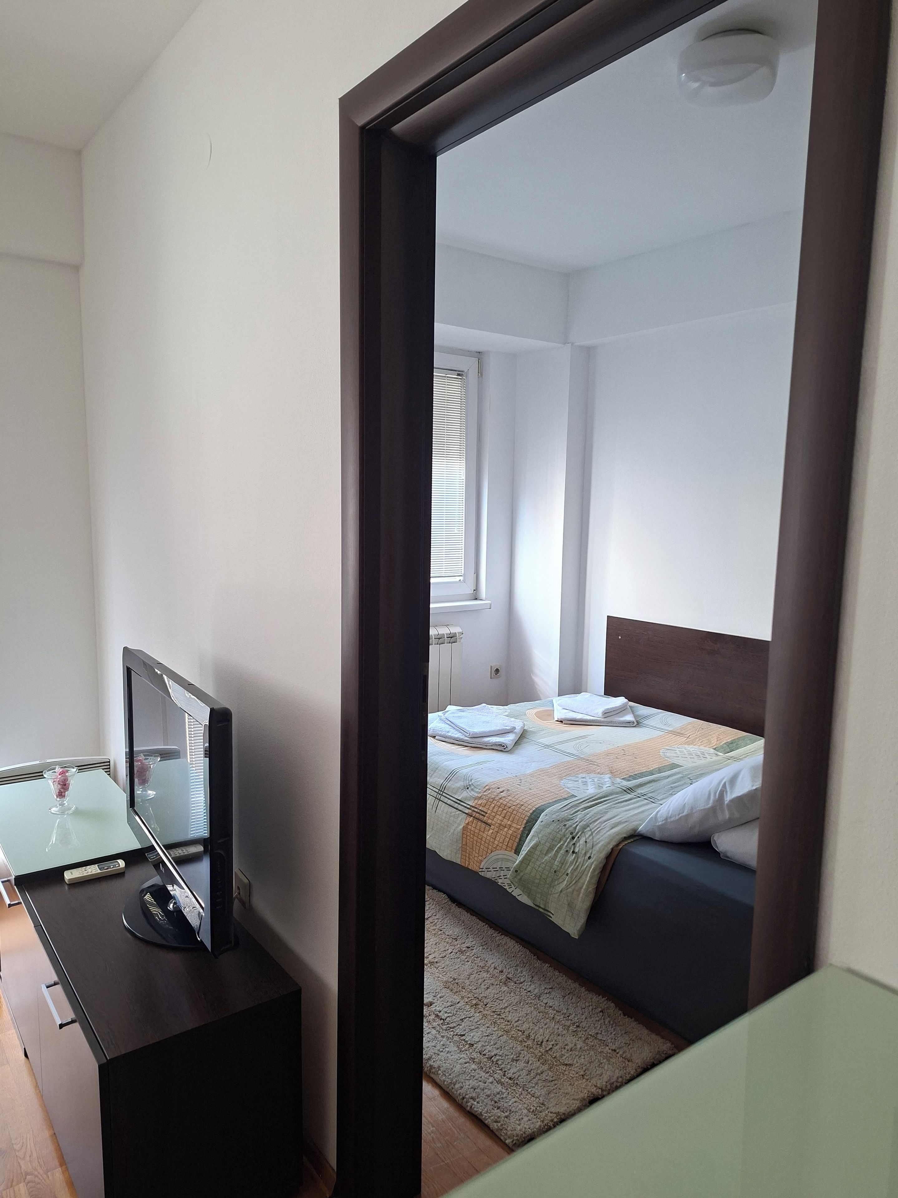 A bedroom with a bed, a bedside table with a television, and a window with blinds.