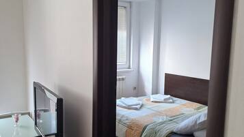 A bedroom with a bed, a bedside table with a television, and a window with blinds.