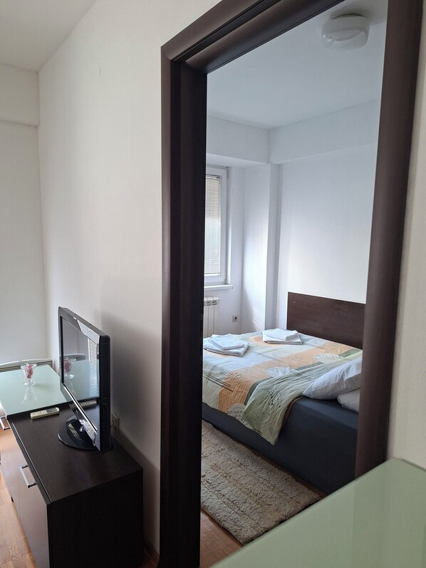 A bedroom with a bed, a bedside table with a television, and a window with blinds. - Fortuna Fortis Apartments - Suite 7 (Skopje)