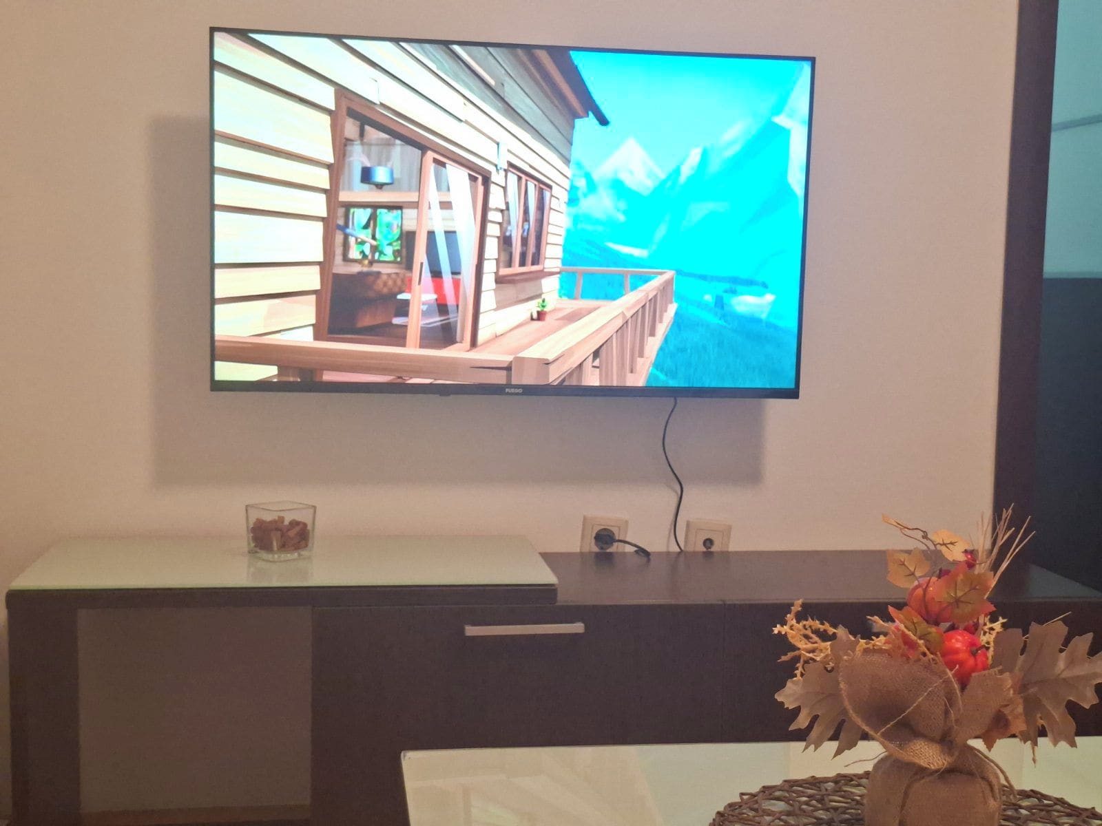 Un televisor montado en la pared, una mesita con un recipiente de vidrio y un arreglo decorativo de plantas.