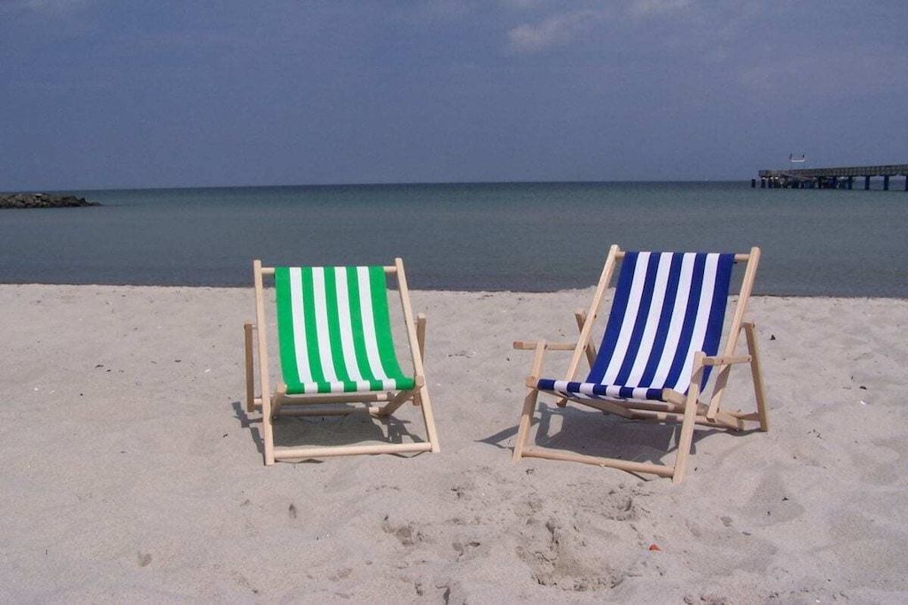 Two beach chairs with striped cushions on a sandy beach with a calm sea and a pier in the background.