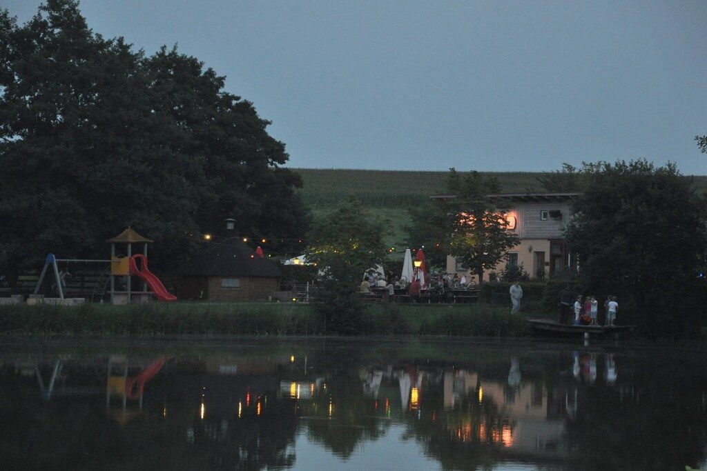 Ein Park am Seeufer mit Spielplatz, einem Gebäude mit Außenbestuhlung und einem Spiegelbild im Wasser.