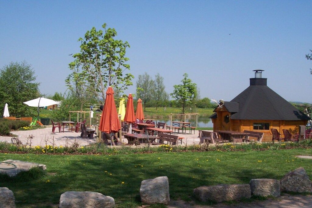 A lakeside outdoor seating area with tables and chairs, a wooden cabin, and a clear sky.