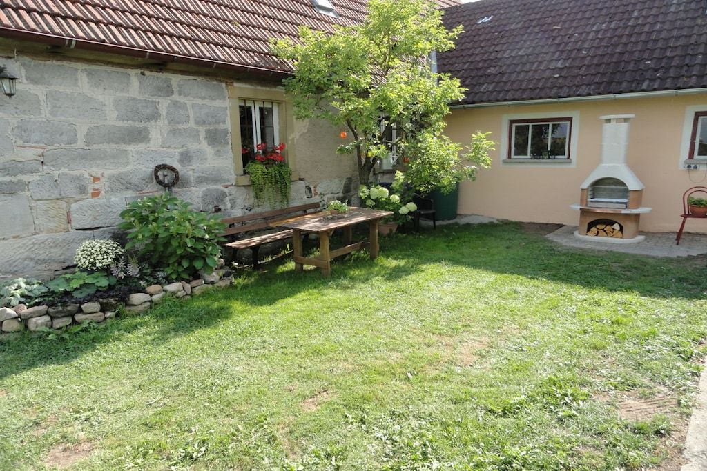 A stone house with a tiled roof, a wooden bench, a small garden with plants, and a wood-fired oven.