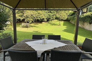 A covered patio with a table and chairs, a glass of water, and a view of a well-maintained lawn and trees. - Villa on Bleidenberg (Oberfell)
