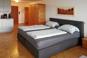 A bedroom with a bed, two pillows, a headboard, a wooden wardrobe, a painting on the wall, and a kitchenette with a coffee maker. - Holiday apartment with 0 bedrooms (Uhldingen-Mühlhofen)