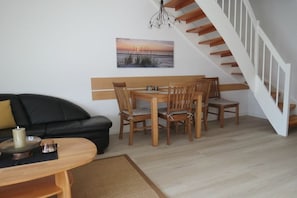 A living room with a black sofa, wooden dining table, and chairs, a staircase, and a painting on the wall. - Seeschlößchen No. 10 (Büsumer Deichhausen)