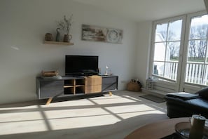 A living room with a flat-screen TV on a dark-colored cabinet, a large window with a view of the outdoors, and a black leather sofa. - Seeschlößchen No. 10 (Büsumer Deichhausen)
