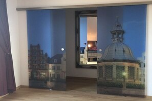 Ein Zimmer mit großem Fenster und Blick auf nächtliche Stadtansicht, an der Wand ein Wandbild eines Gebäudes.