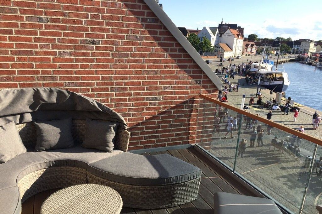 Ein Balkon mit Korbmöbeln, einer Ziegelwand und Blick auf ein Ufergebiet mit Menschen und Booten.