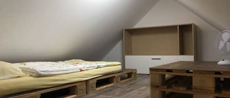 A small, single-room apartment with a loft bed, a wooden desk, and a built-in shelving unit.