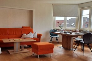 A living room with a wooden coffee table, orange sofa, and a dining area with a wooden table and chairs. - Holiday apartment in Zingst with balcony (Zingst)