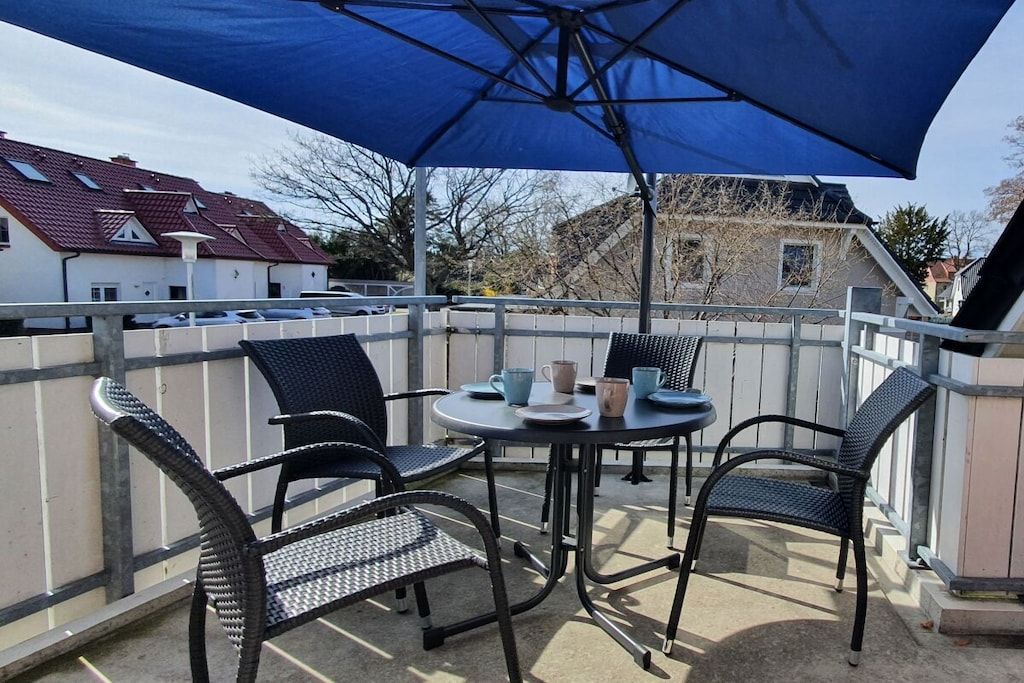 A balcony with a table and chairs, a blue umbrella, and a view of houses in the background.