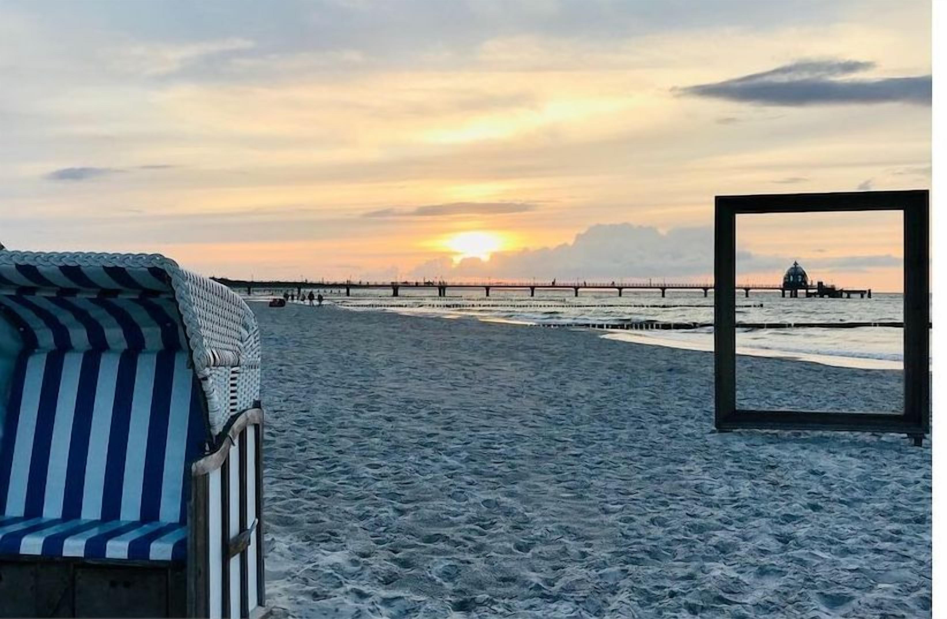 A beach scene with a chair and a framed picture, a pier in the background, and a sunset.