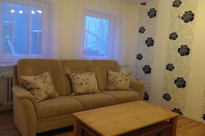 Ein Wohnzimmer mit einem beigefarbenen Sofa, zwei passende Kissen, einem Holztisch und einem Fenster mit Blick auf kahle Bäume.