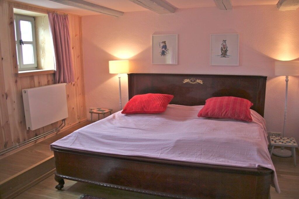 Ein Schlafzimmer mit einem hölzernen Kopfteil, zwei Nachttischlampen, einem Fenster mit Vorhängen und zwei gerahmten Bildern an der Wand.