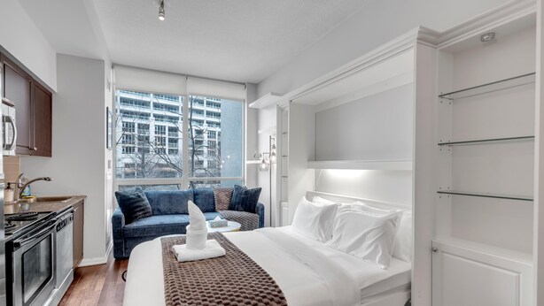 A modern hotel room with a bed, a sofa, a kitchen area, and a large window with a city view. - StayToronto - Toronto Central Studio - DT (Toronto)