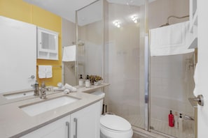 Une salle de bain avec une douche à parois de verre, un lavabo blanc et un mur d’accent jaune.